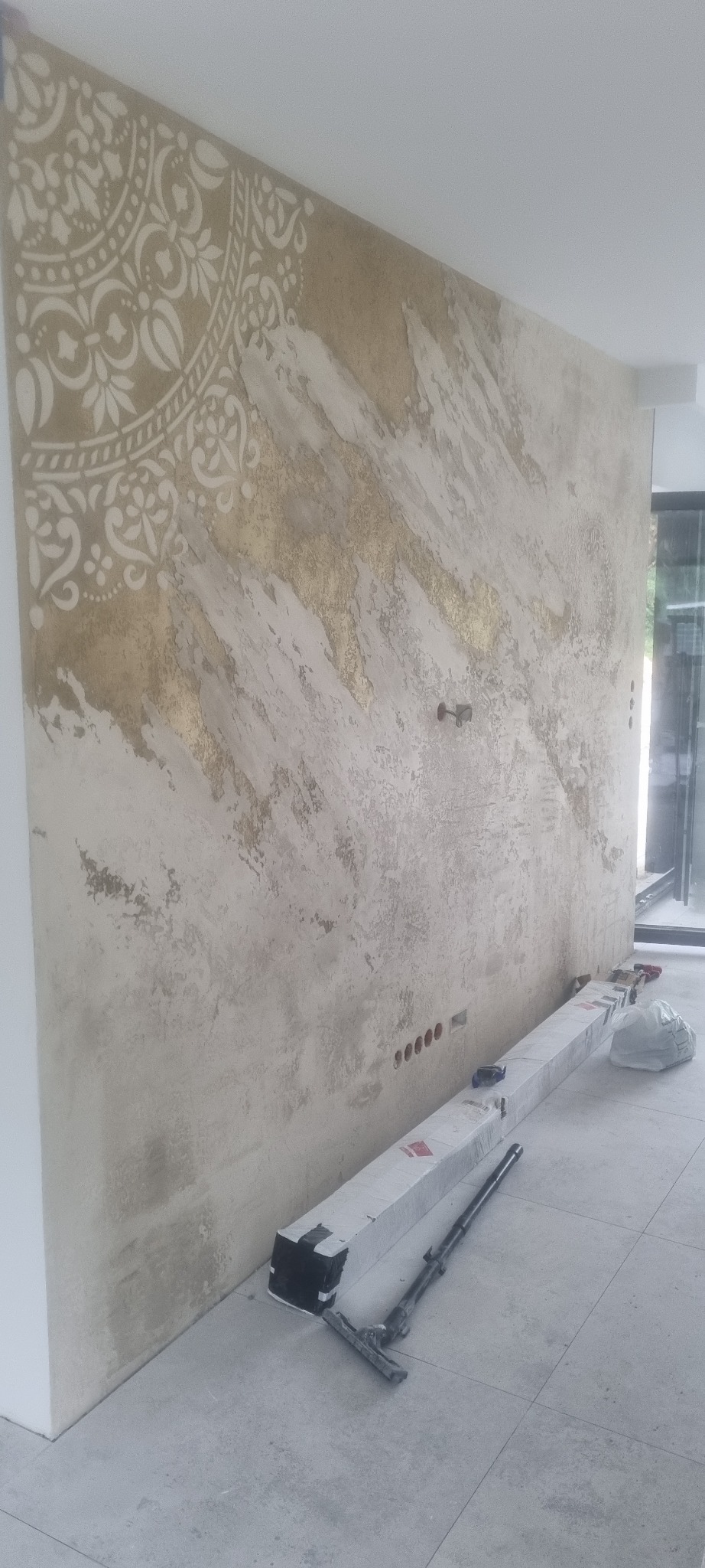 Dekoracyjna ściana z efektem stiuku w odcieniach beżu i złota, zdobiona szablonowym wzorem przypominającym mandalę, z widocznymi puszkami elektrycznymi i narzędziami malarskimi w tle.