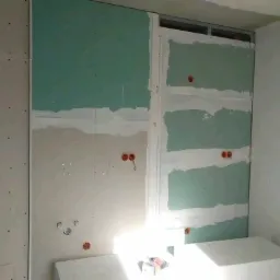 Montaż płyt gipsowo-kartonowych na ścianie z widocznymi puszkami elektrycznymi i częściowo zaszpachlowanymi łączeniami, oświetlenie od dołu.