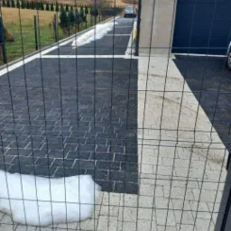 Nowoczesna posesja z czarno-białą kostką brukową, widoczna przez metalowe ogrodzenie, z resztkami śniegu przy krawędzi.