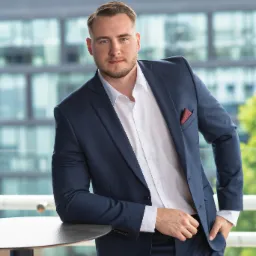 Młody mężczyzna w granatowym garniturze i białej koszuli, oparty o stół na tle nowoczesnego biurowca. Widoczny fragment czerwonej poszetki w kieszonce marynarki.