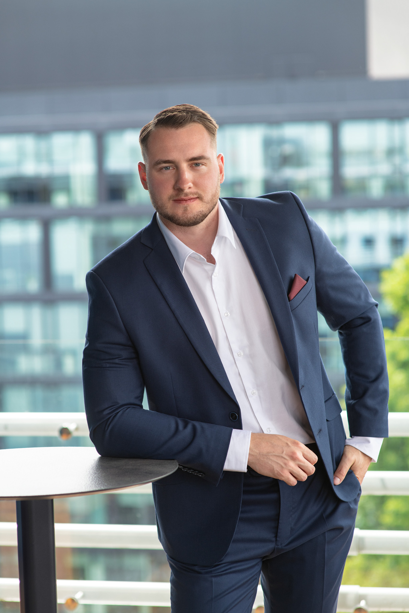 Młody mężczyzna w granatowym garniturze i białej koszuli, oparty o stół na tle nowoczesnego biurowca. Widoczny fragment czerwonej poszetki w kieszonce marynarki.