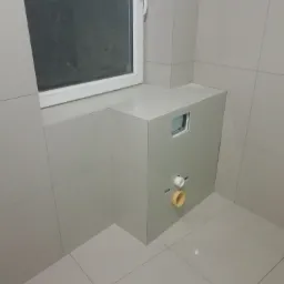 Szara łazienka w trakcie remontu, widoczna zabudowa stelaża WC z wystającymi rurami kanalizacyjnymi, położone płytki na podłodze i ścianach, okno w tle.