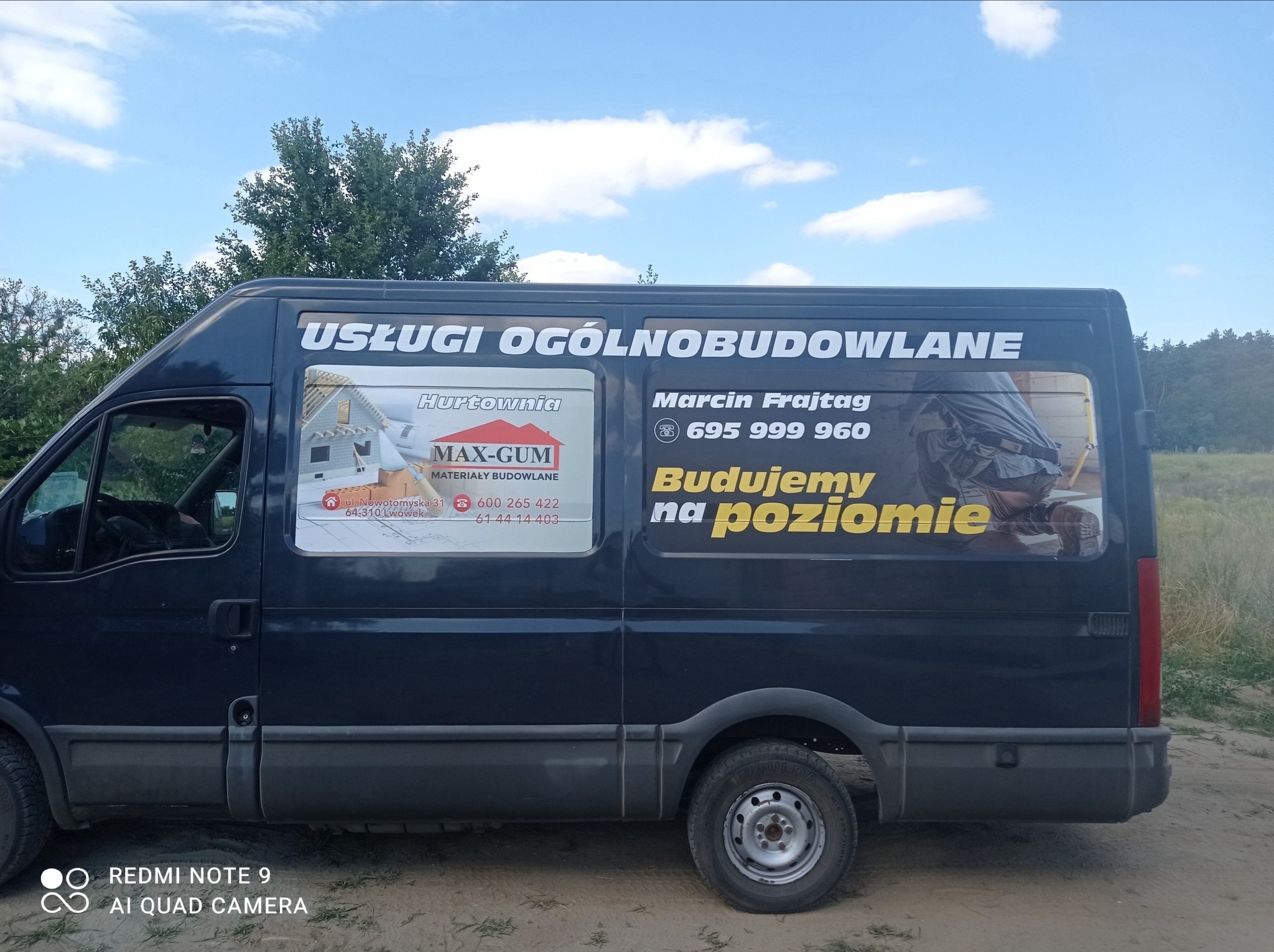 Ciemny bus z reklamą usług ogólnobudowlanych, logo Max-Gum, numerem telefonu i hasłem 'Budujemy na poziomie', zaparkowany na piaszczystej drodze na tle drzew i błękitnego nieba.