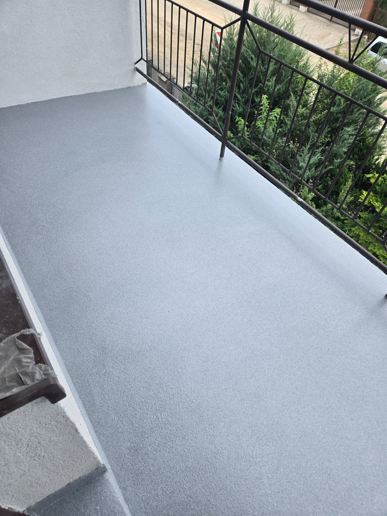 Szara, gładka powierzchnia balkonu z metalową barierką i widokiem na zieleń. Nowoczesne wykończenie, równa i jednolita struktura posadzki.