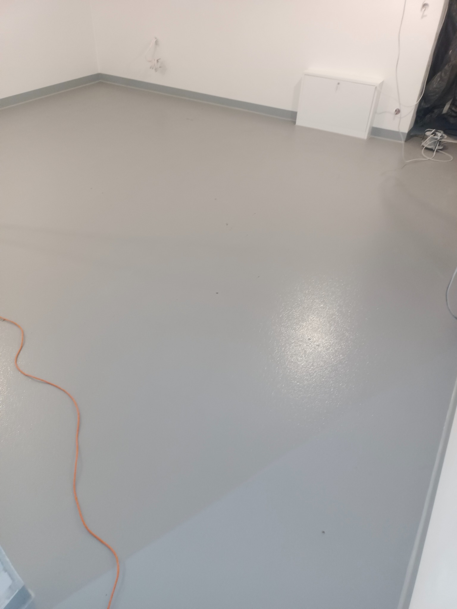 Szara, gładka posadzka żywiczna w jasnym pomieszczeniu. Widoczne listwy przypodłogowe i fragment przewodu. Minimalistyczne wykończenie wnętrza z bliska.