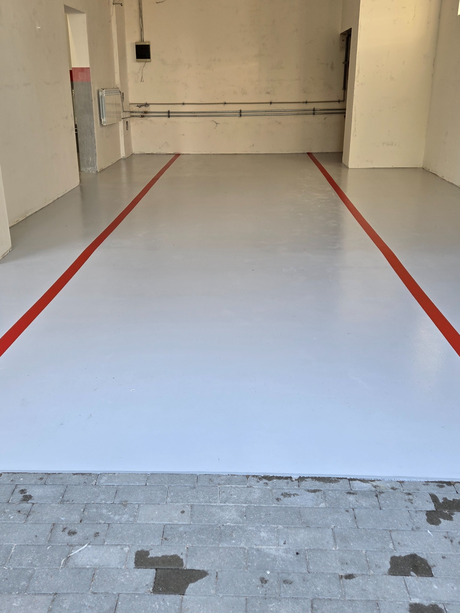 Jasnoszara, gładka podłoga z czerwoną linią wyznaczającą pole wewnątrz pomieszczenia. Widoczne ściany i fragment posadzki z kostki brukowej przy wejściu.