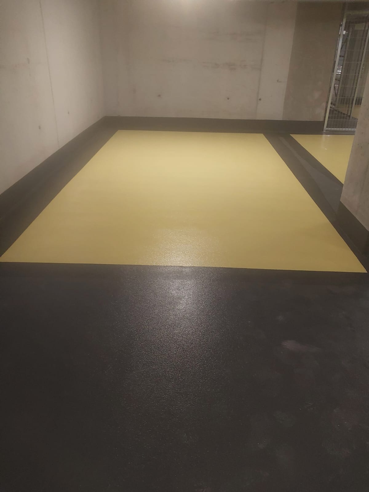 Żółto-czarna, gładka posadzka w surowym wnętrzu. Widoczny fragment ściany i metalowa krata w tle. Podłoga podzielona na strefy kolorystyczne.