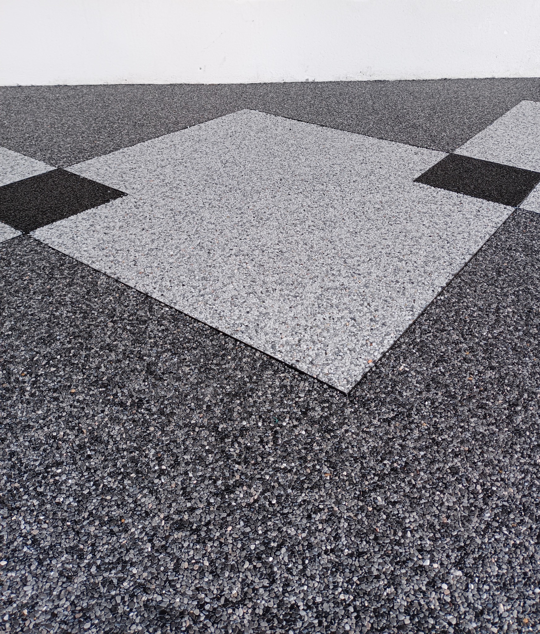 Dekoracyjna, dwukolorowa posadzka z kamyczków żywicznych ułożona w szachownicę. Szara i czarna mozaika na podłodze, biała ściana w tle.