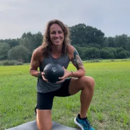 Uśmiechnięta kobieta w sportowym stroju klęczy na macie na trawie, trzymając czarną piłkę lekarską z napisem '12 LB', widoczne drzewa w tle.