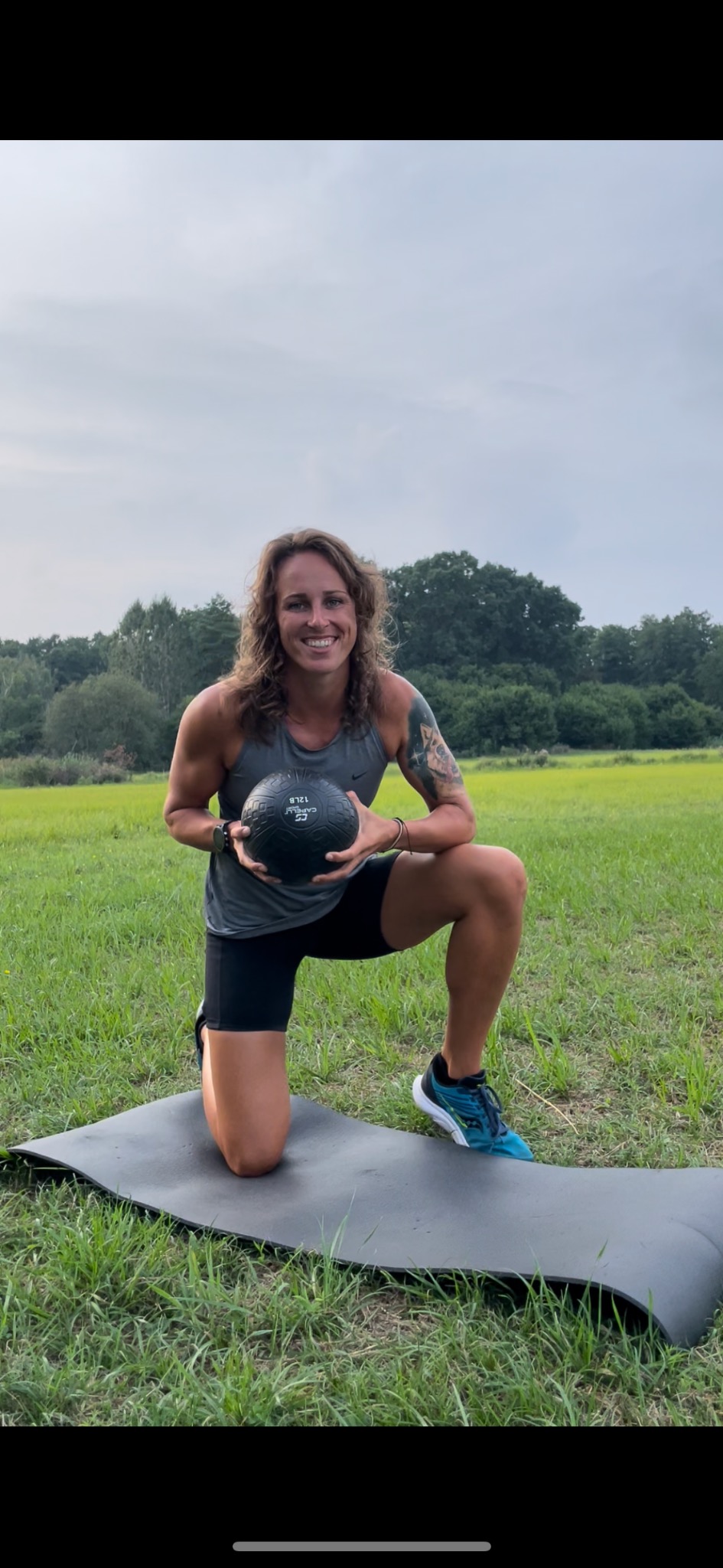 Uśmiechnięta kobieta w sportowym stroju klęczy na macie na trawie, trzymając czarną piłkę lekarską z napisem '12 LB', widoczne drzewa w tle.