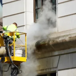 Pracownik w żółtym kombinezonie na podnośniku koszowym czyści elewację budynku z kamienia za pomocą myjki ciśnieniowej, widoczna para wodna i brud spływający po ścianie.