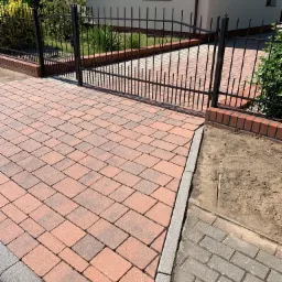 Nowo ułożona, czerwono-szara kostka brukowa prowadząca do czarnej, metalowej bramki i ogrodzenia na tle jasnego domu z ciemnymi oknami.