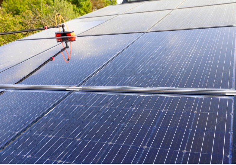 Szczotka na wysięgniku czyści panele słoneczne na dachu, widoczne drzewa w tle.