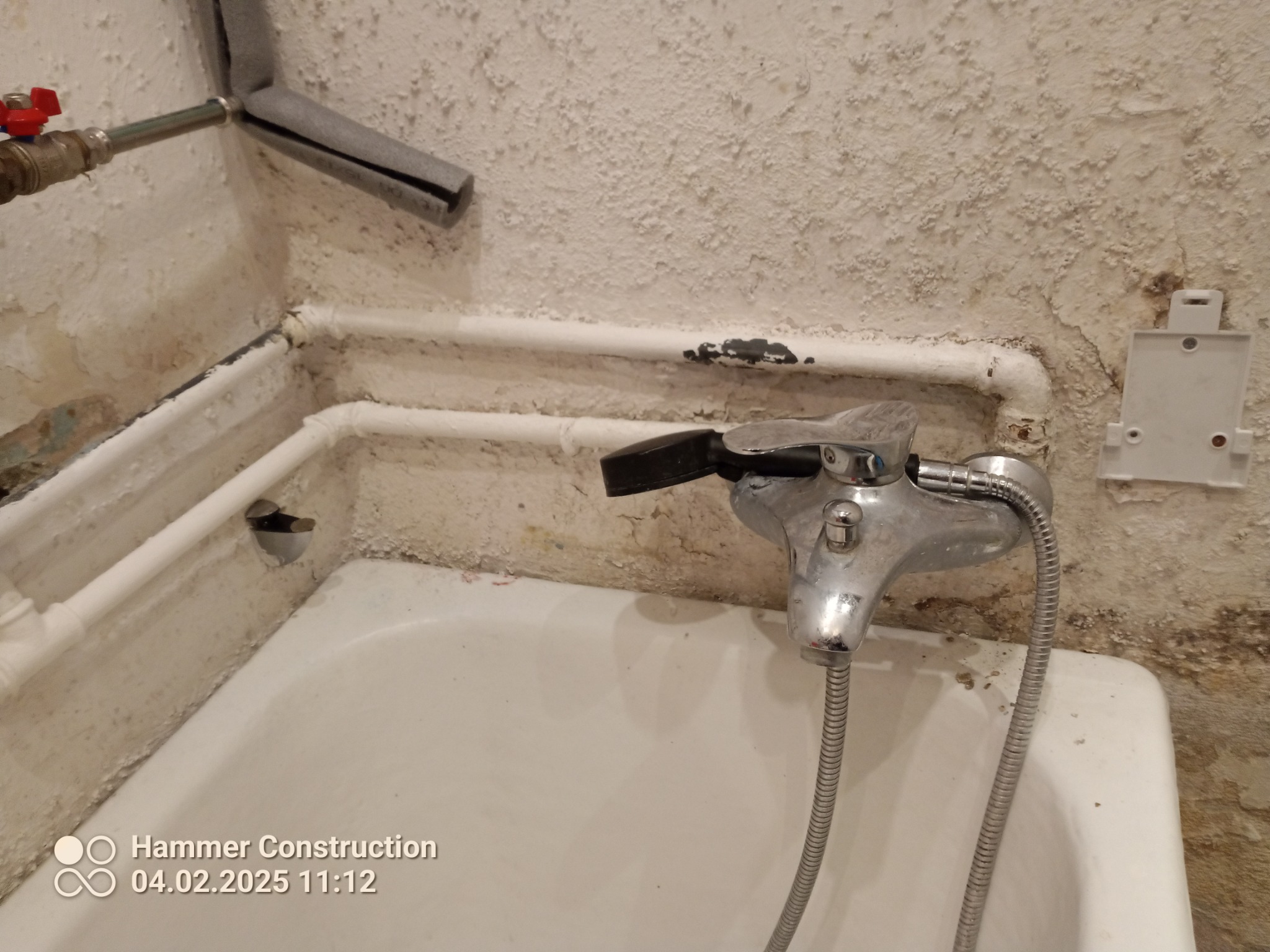 Stara, biała wanna z zamontowaną baterią z wężem, widoczne zniszczone ściany z odpryskującą farbą i rurami instalacji wodnej.