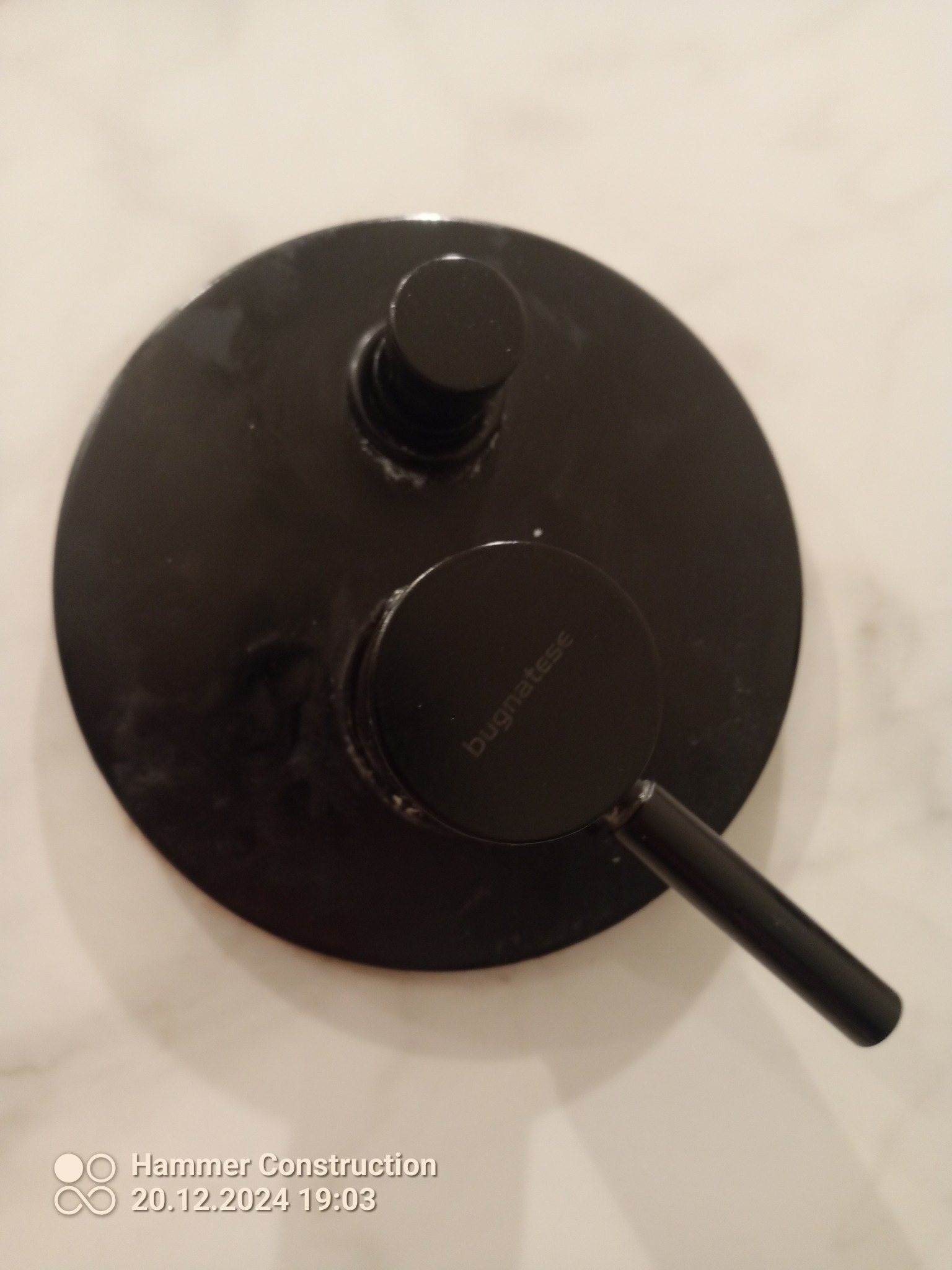 Czarny, okrągły zawór termostatyczny marki Bugnatese zamontowany na jasnej, marmurowej ścianie w łazience, widoczne ślady montażu.