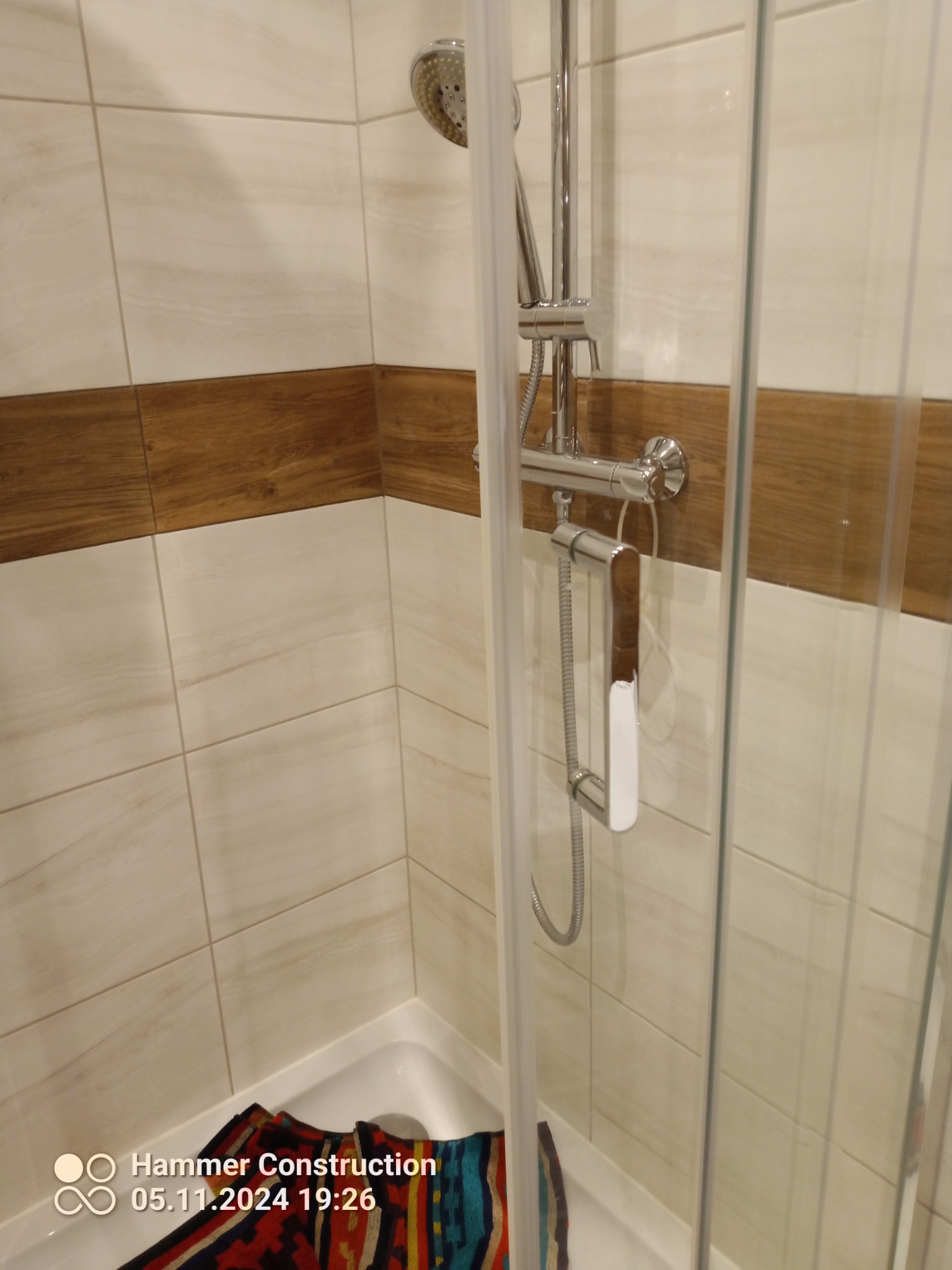 Nowoczesna kabina prysznicowa z chromowaną armaturą i beżowo-brązowymi płytkami imitującymi drewno, widoczne detale wykończenia i kolorowy ręcznik w brodziku.