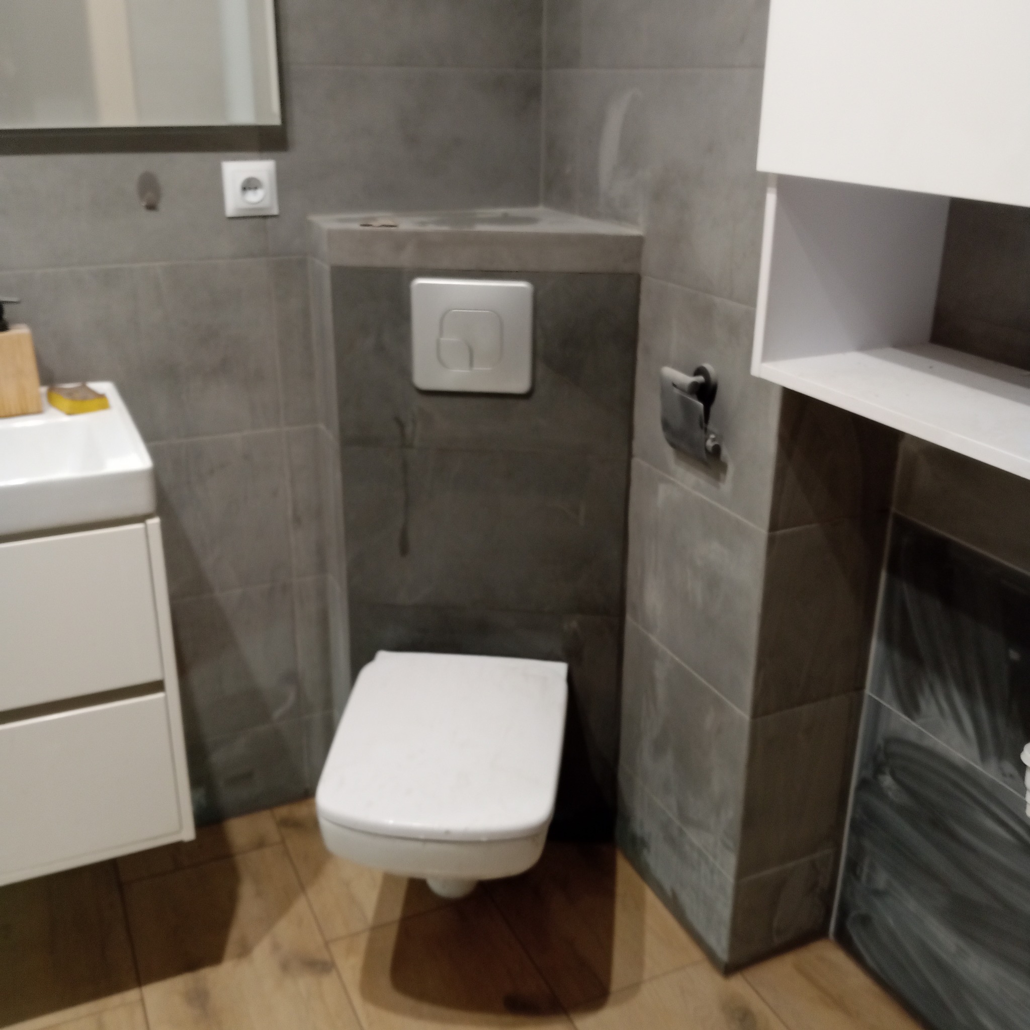 Nowoczesna, szara łazienka z podwieszaną toaletą, umywalką z szafką i drewnopodobną podłogą. Widoczny fragment lustra, biała szafka wisząca i przycisk spłuczki.