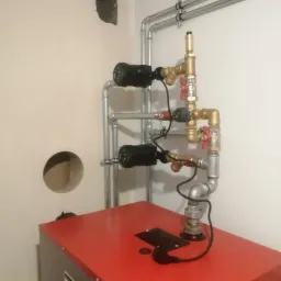 Czerwony piec grzewczy Defro z podłączonymi rurami i zaworami, widoczne dwa czarne pompy obiegowe, fragment ściany z otworem i uszkodzonym tynkiem.