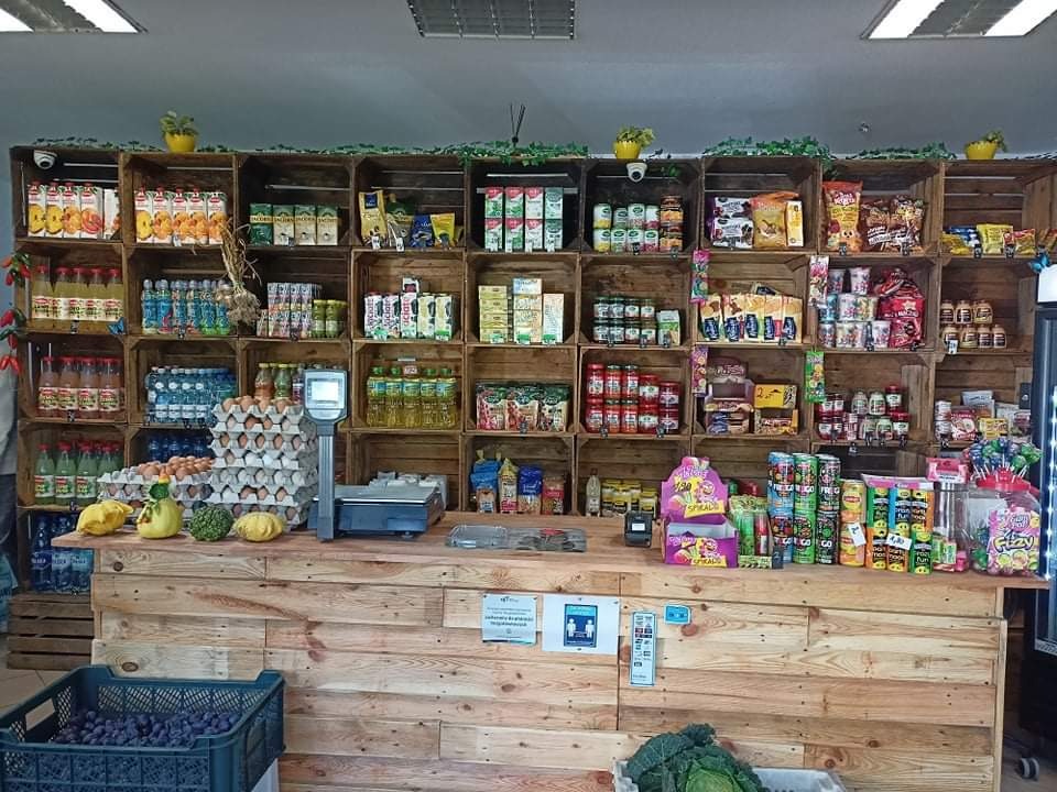 Kącik sklepowy z drewnianymi skrzynkami wypełnionymi produktami spożywczymi, w tle zielona dekoracja roślinna, na pierwszym planie lada z drewnianych desek i waga sklepowa.