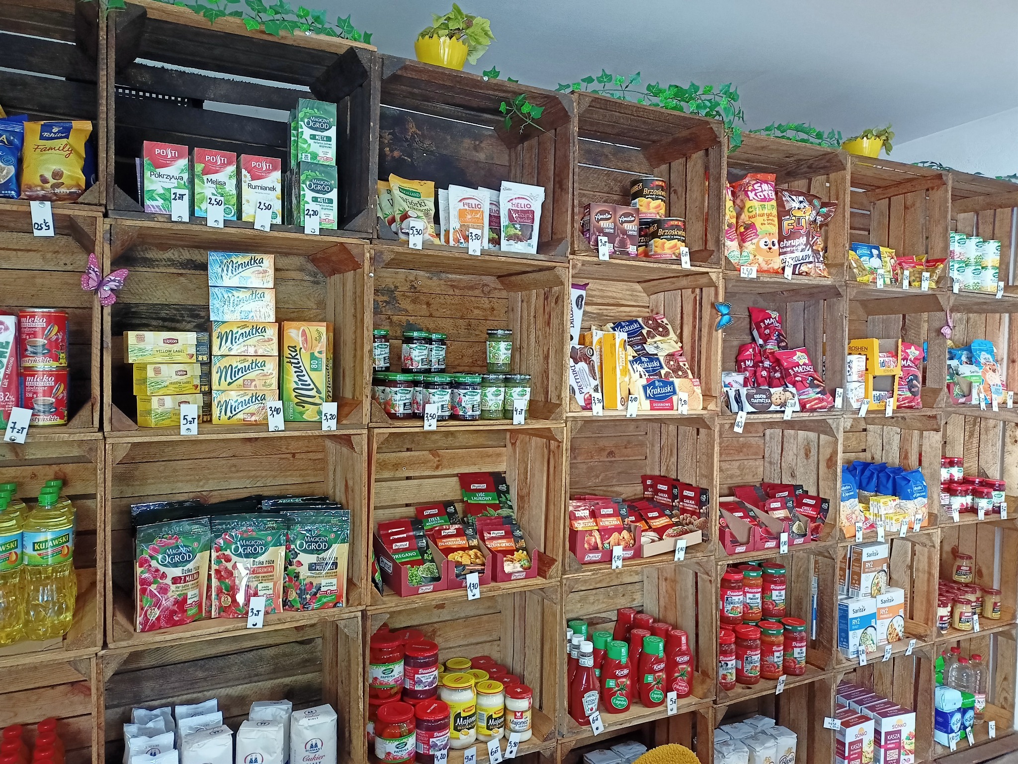 Ekspozycja produktów spożywczych w sklepie, prezentacja w drewnianych skrzynkach, różnorodność artykułów z cenami.