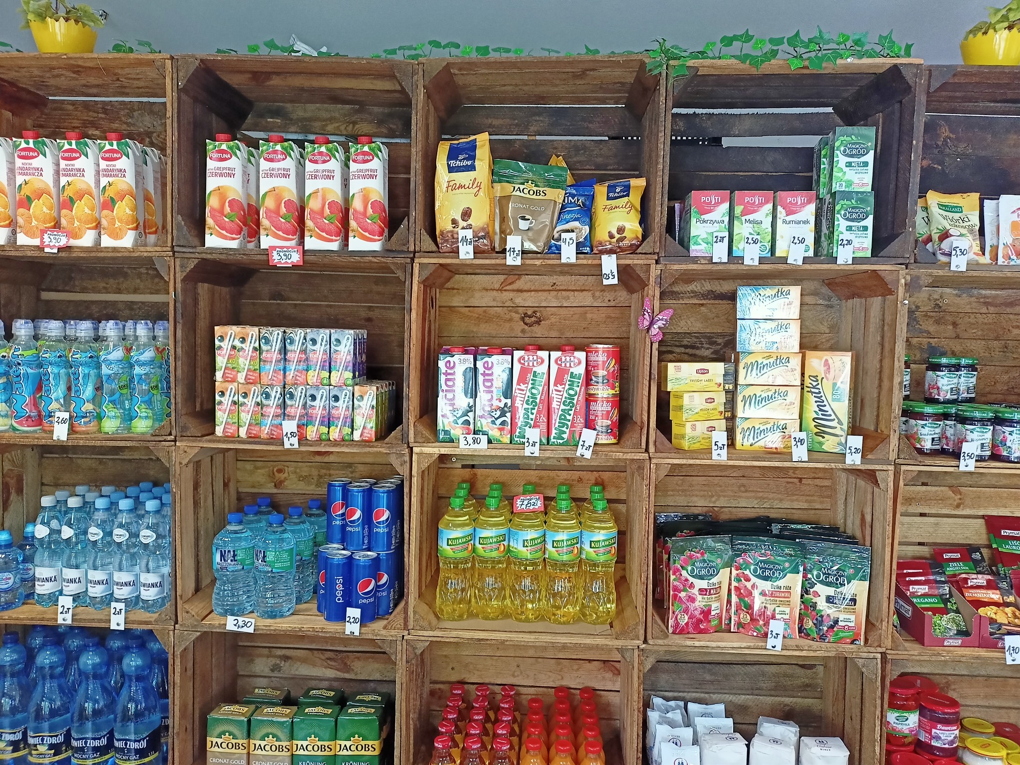 Regał sklepowy z drewnianych skrzynek wypełniony różnorodnymi produktami spożywczymi, napojami, kawami, herbatami, olejami i przyprawami, z cenami umieszczonymi przy każdym produkcie.