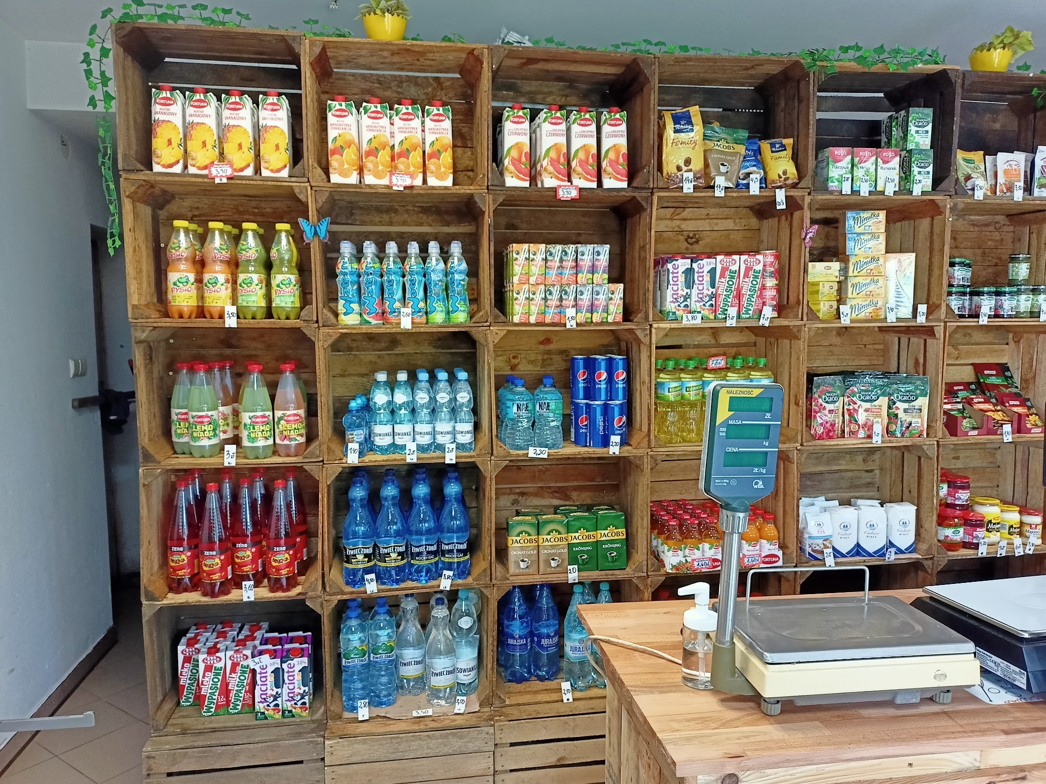 Półki z drewnianych skrzynek wypełnione różnorodnymi napojami, wodami, sokami i kawami w sklepie w Łodzi, obok waga sklepowa.