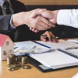 Uścisk dłoni nad umową, obok model domu, plik banknotów i monety na biurku.