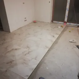 Układanie płytek podłogowych o wzorze marmuru w trakcie prac, widoczne narzędzia glazurnicze, poziomica laserowa, fragment surowej podłogi obok ułożonych płytek.