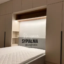 Sypialnia z łóżkiem i zabudową meblową na wymiar w odcieniach beżu i bieli, z podświetlaną wnęką nad wezgłowiem i czarnymi, minimalistycznymi uchwytami szaf.