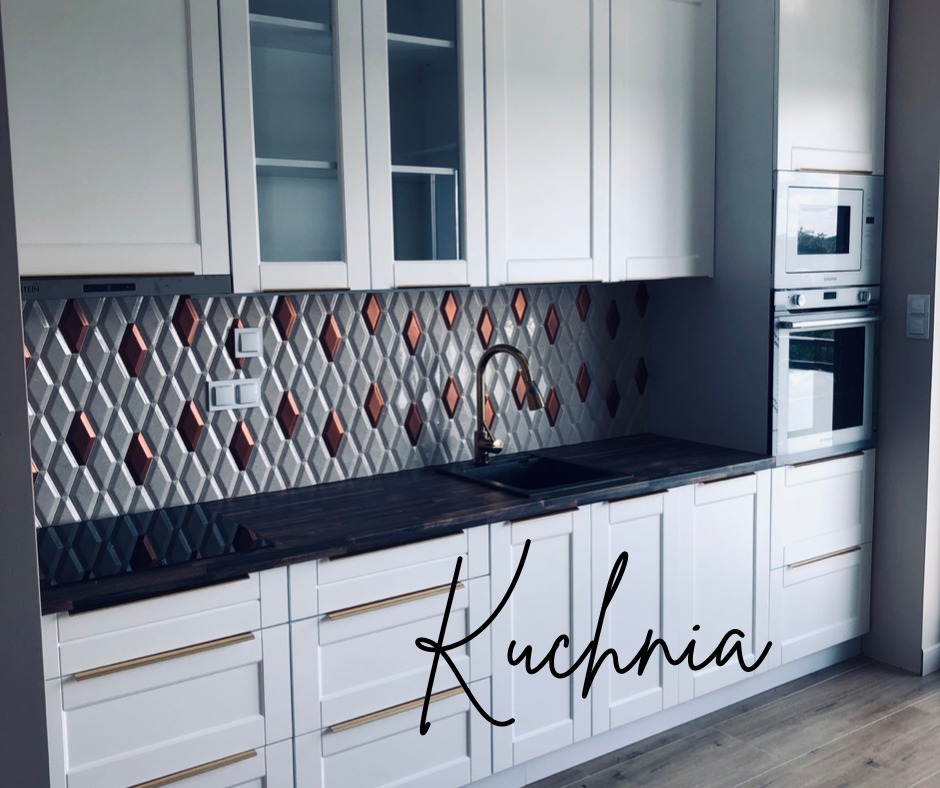Nowoczesna kuchnia z białymi frontami szafek, ciemnym blatem i geometryczną mozaiką w odcieniach szarości i miedzi na ścianie nad blatem, złote uchwyty, zabudowany piekarnik i mikrofalówka.