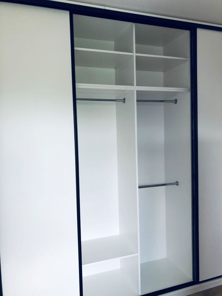 Wnętrze białej szafy przesuwnej z czarnymi ramami, widoczne półki i drążki na ubrania, jedna z częściowo otwartych drzwi.