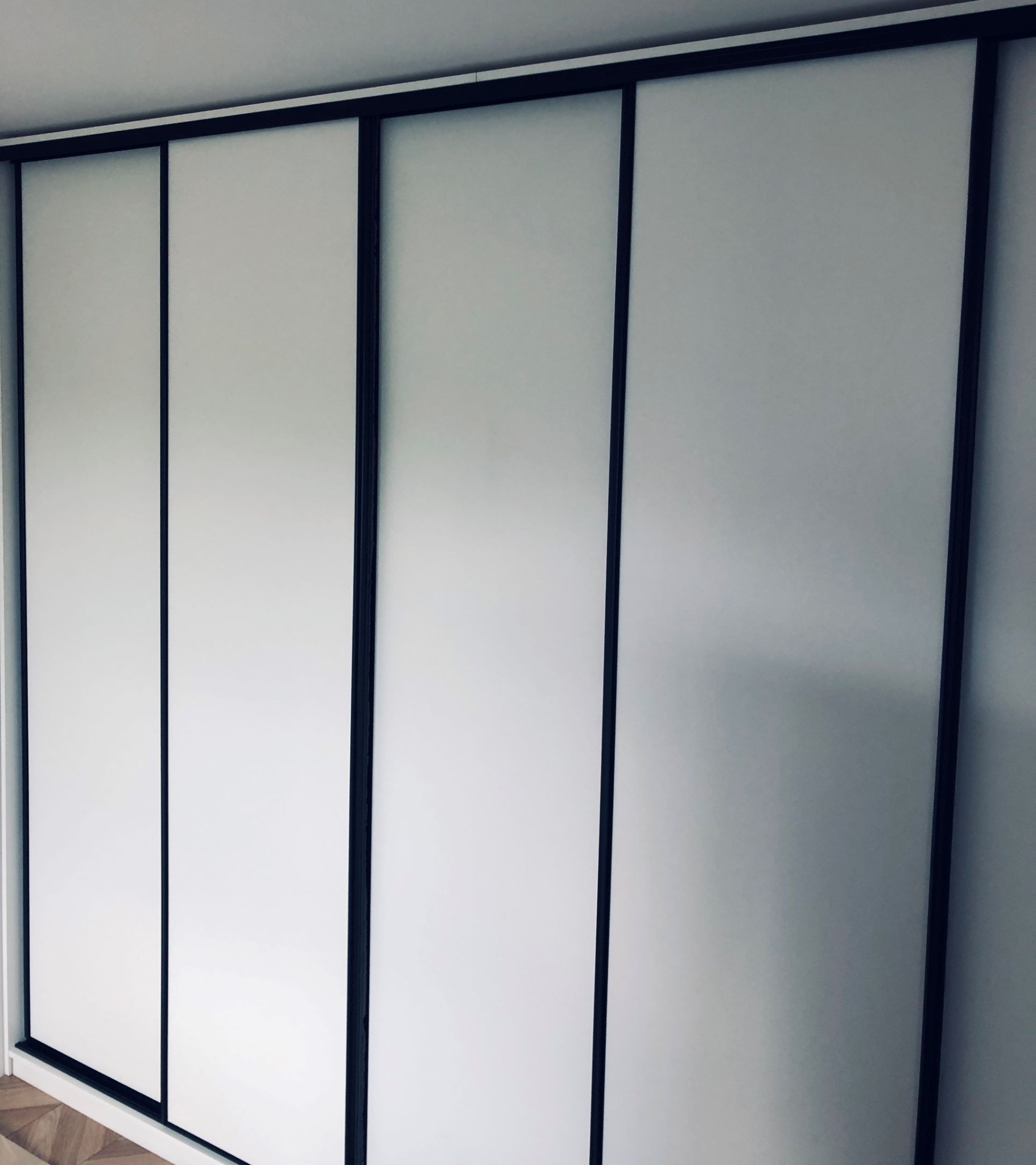 Minimalistyczna szafa przesuwna z białymi frontami i ciemnymi, wąskimi ramami, widoczna z perspektywy podłogi z parkietem ułożonym we wzór jodełki.