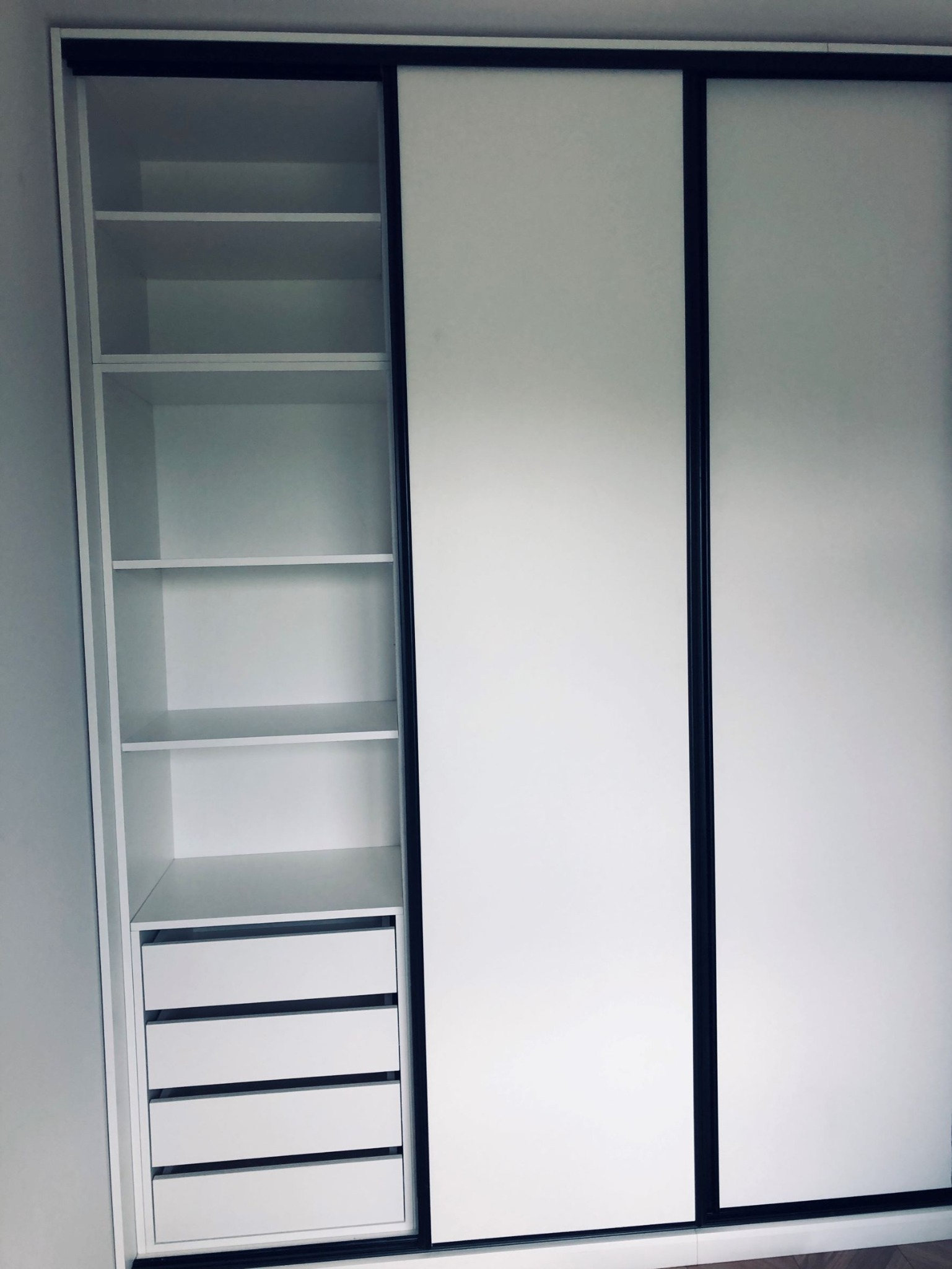 Biała szafa wnękowa z przesuwnymi drzwiami i czarnymi obramowaniami, z widocznymi półkami i szufladami po lewej stronie.