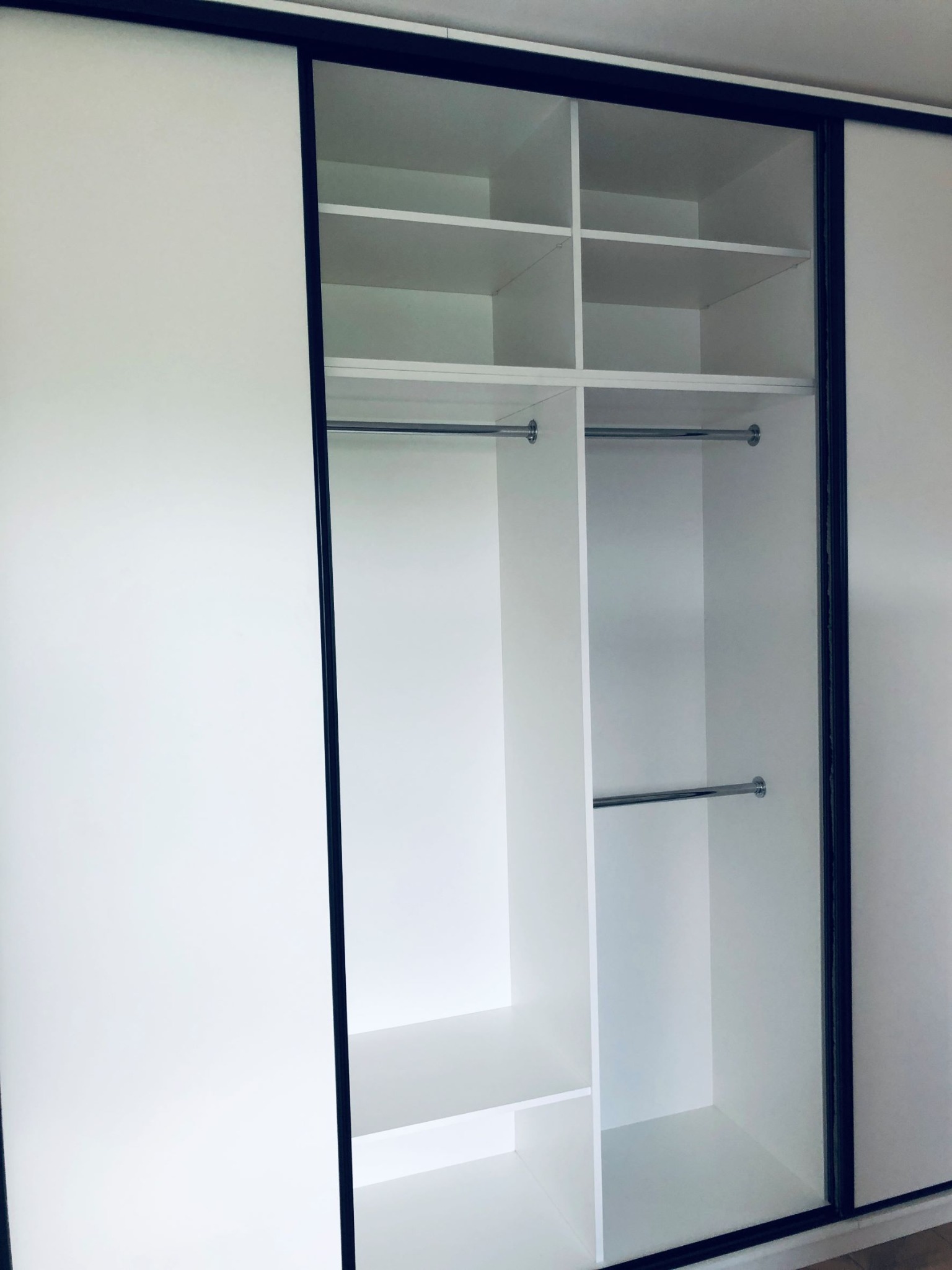 Wnętrze białej szafy przesuwnej z czarnymi obramowaniami, widoczne półki i drążki na ubrania, drzwi częściowo zasłonięte.