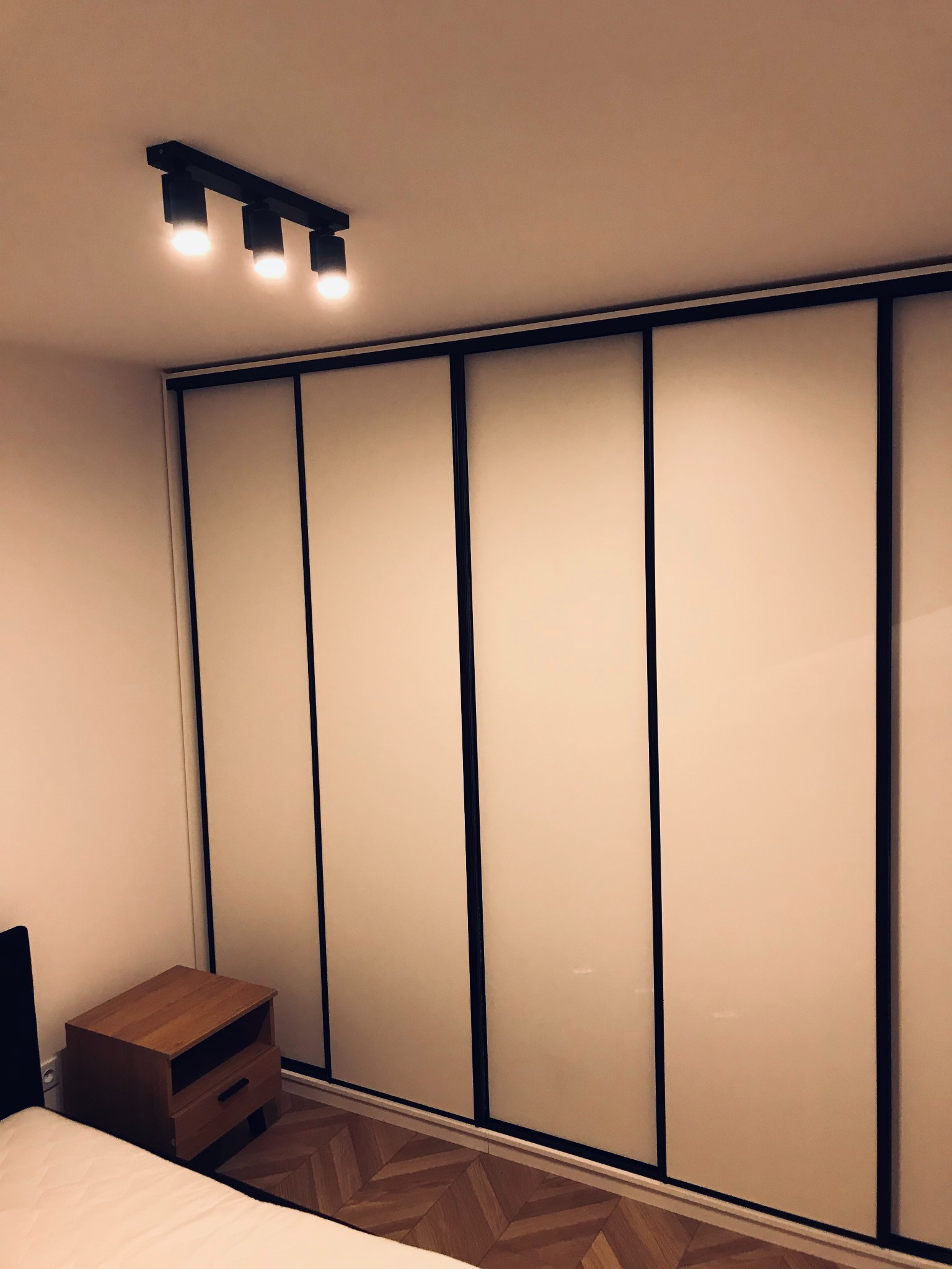 Wnętrze sypialni z dużą szafą wnękową z przesuwnymi, jasnymi drzwiami z czarnymi ramami i drewnianą szafką nocną obok łóżka, oświetloną trzypunktową lampą sufitową.