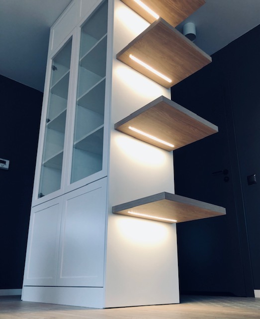 Biała zabudowa meblowa z przeszkloną witryną i trzema podświetlanymi półkami z drewna, minimalistyczny design, ciemne ściany w tle.