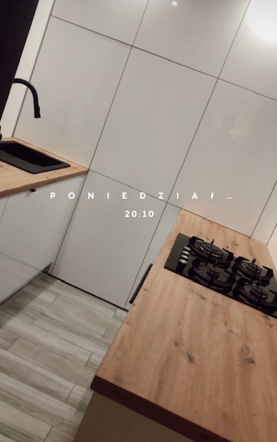 Nowoczesna kuchnia z białymi, gładkimi frontami, drewnianym blatem i czarną armaturą, widok z góry lekko zniekształcony przez filtr, z widocznym fragmentem płyty gazowej i zlewu.