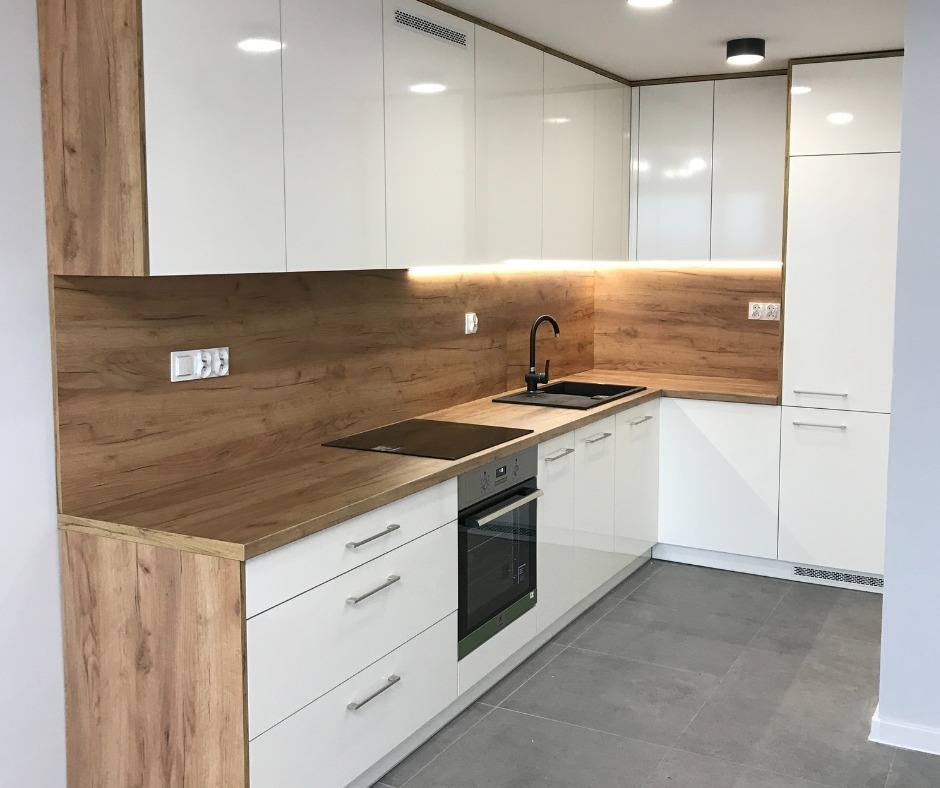 Nowoczesna kuchnia z białymi, błyszczącymi frontami szafek, drewnianym blatem i panelem nad blatem w kolorze drewna, czarną armaturą i szarą podłogą. Oświetlenie podszafkowe LED podkreśla ciepły...