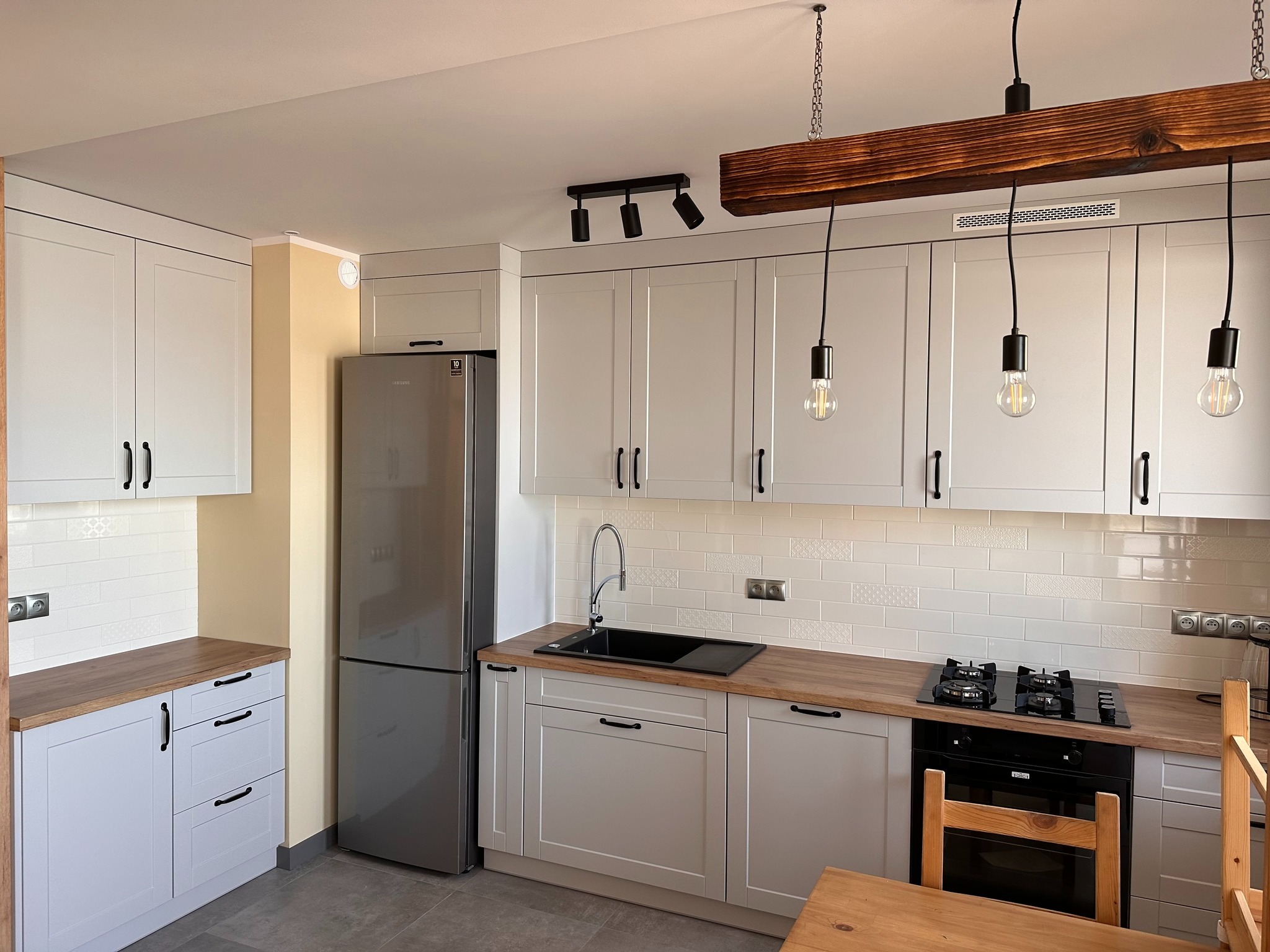 Jasna kuchnia z drewnianymi blatami i czarnymi uchwytami, oświetlona drewnianą belką z trzema wiszącymi żarówkami, widoczna płyta gazowa i zlewozmywak.