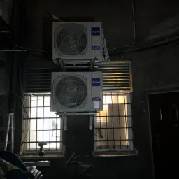 Dwa klimatyzatory Haier zamontowane jeden nad drugim na elewacji budynku, widoczne okna z kratami i wiertarka na parapecie, słabe oświetlenie