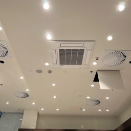 Smart Free Wind - Sufit w Lesznie z zamontowaną kasetą klimatyzacyjną, anemostatami i punktowymi lampami LED. Widoczna drabina świadczy o trwających pracach instalacyjnych.