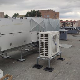 Smart Free Wind - Biała jednostka zewnętrzna klimatyzacji zamontowana na metalowej konstrukcji z gumowymi podkładkami na dachu budynku, obok widoczny kanał wentylacyjny i skrzynka z narzędziami.