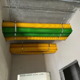 Żółto-zielone rury wentylacyjne karbowane, zamocowane do sufitu za pomocą metalowych obejm, widoczne w surowym wnętrzu budynku w trakcie wykończenia.