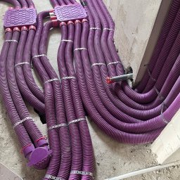 Smart Free Wind - Fioletowe rury wentylacyjne karbowane prowadzone od rozdzielaczy, zamocowane metalowymi opaskami na surowej podłodze w trakcie montażu systemu rekuperacji.