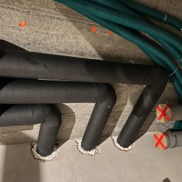 Smart Free Wind - Czarny system rur izolowanych termicznie, wychodzących ze ściany i podłogi, uszczelnionych pianką montażową, obok zielonych rur wentylacyjnych przymocowanych do sufitu, na surowym betonowym tle.