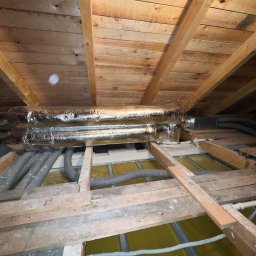 Smart Free Wind - Izolowane rury wentylacyjne i elastyczne przewody w przestrzeni poddasza, widoczne drewniane belki stropowe i jasne panele izolacyjne.