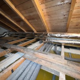 Smart Free Wind - Szare, karbowane rury wentylacyjne i srebrne, zaizolowane przewody rekuperacji zamontowane pod drewnianą konstrukcją dachu na poddaszu w trakcie instalacji.