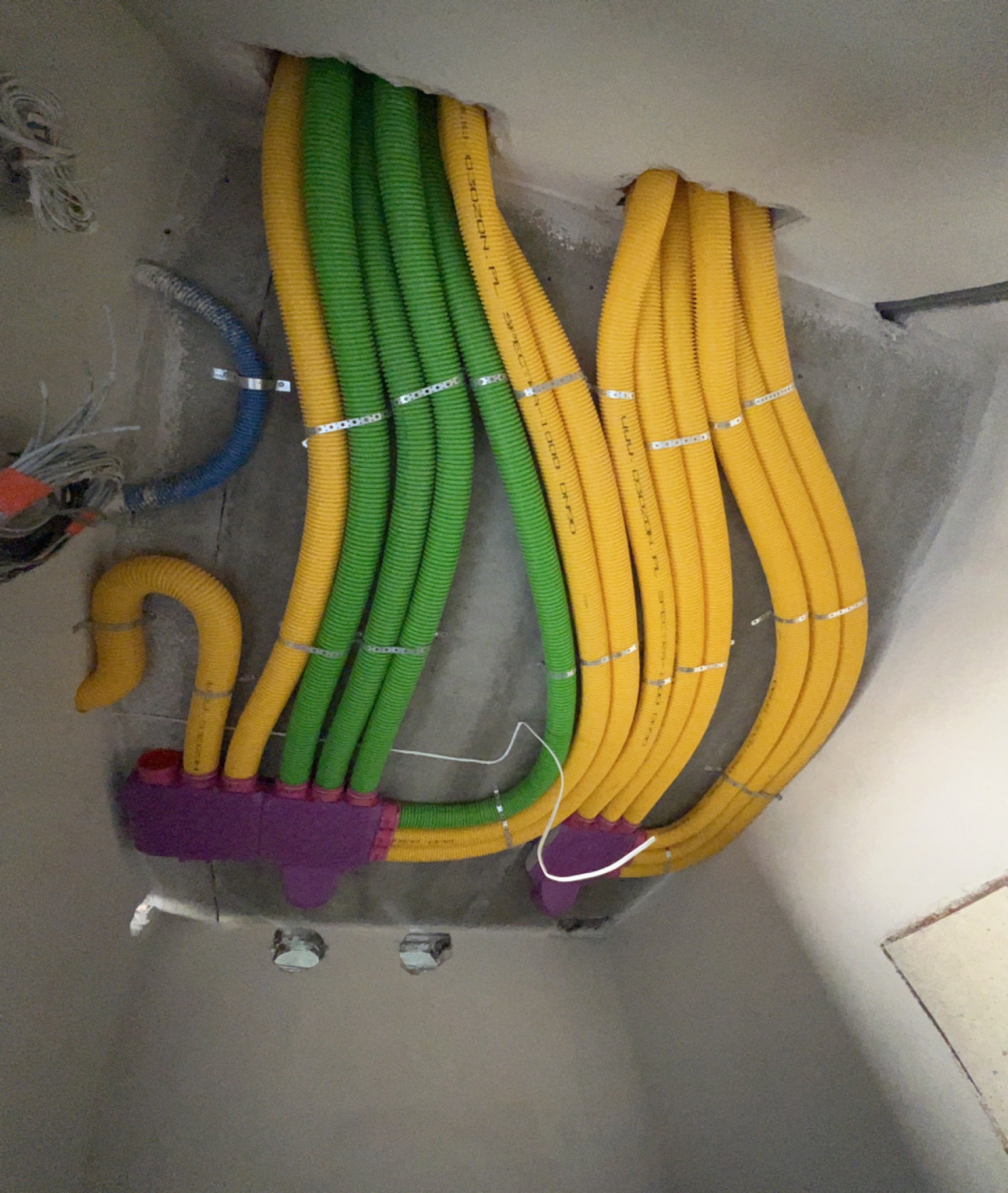 Żółte i zielone karbowane rury wentylacyjne przymocowane metalowymi obejmami do ściany, połączone fioletową skrzynką rozdzielczą, widoczne otwory w ścianie.