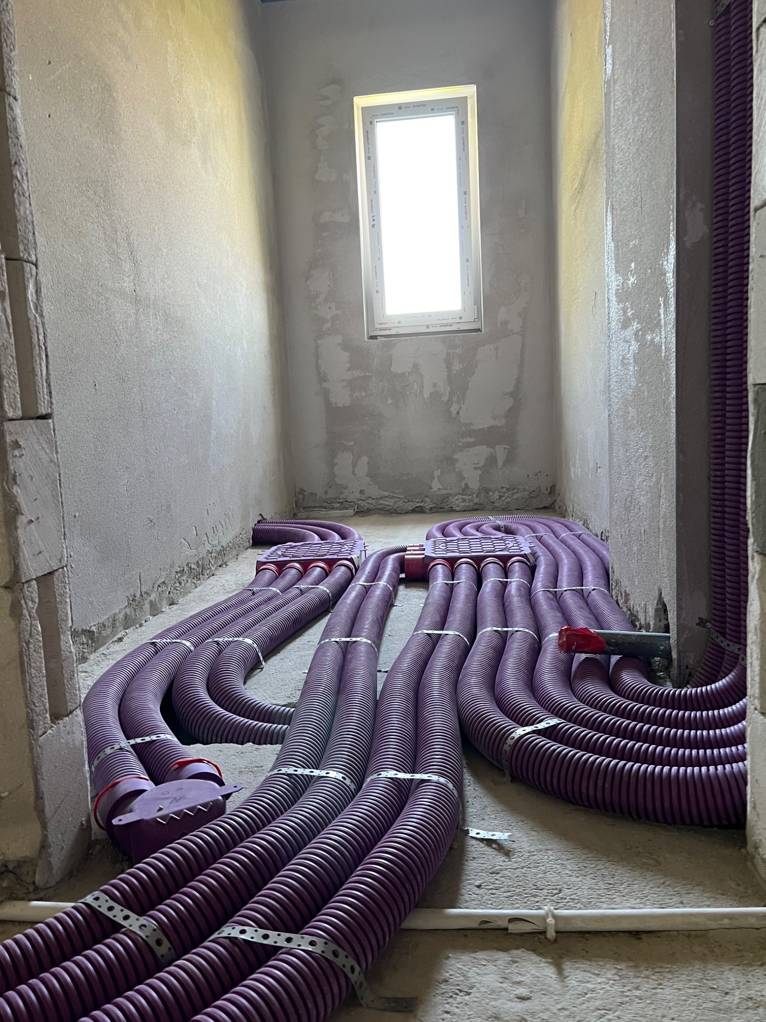 Fioletowe rury wentylacyjne ułożone na surowej podłodze w trakcie montażu w pomieszczeniu z oknem i surowymi ścianami.