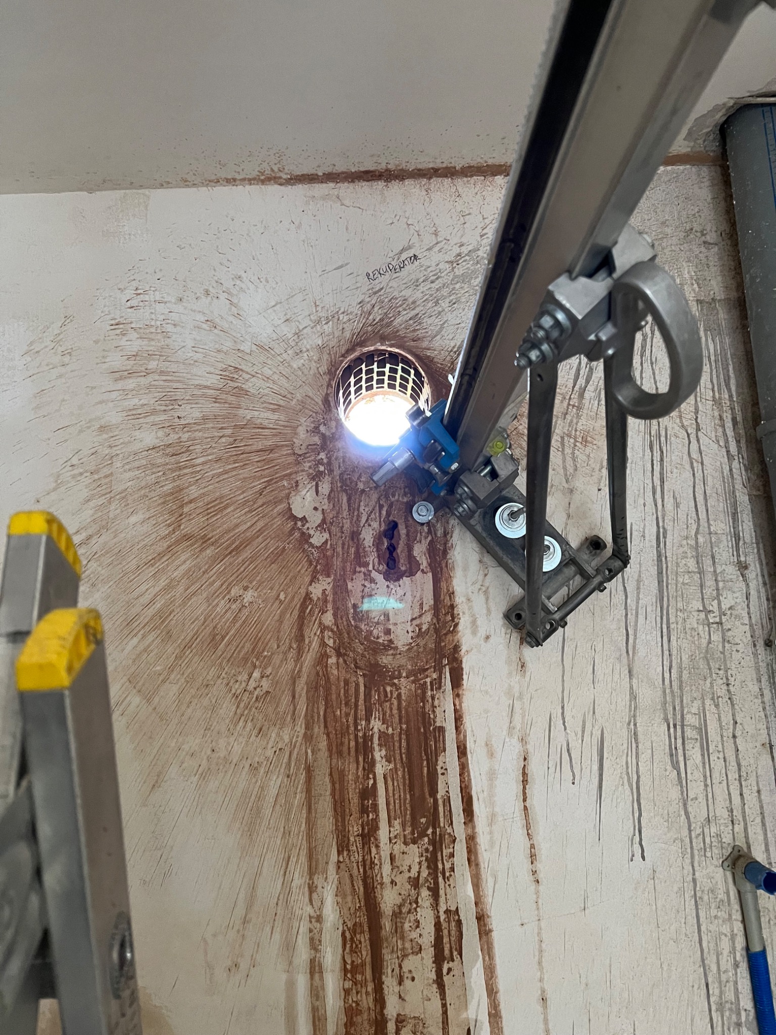 Wiercenie otworu w ścianie pod instalację wentylacyjną, widoczne ślady po wierceniu i metalowa konstrukcja wiertła z oświetleniem.